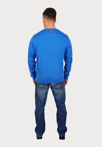 Kungligblå sweatshirt med långa ärmar och ribbade muddar, kombinerad med ljusblå jeans. Märkbar röd och vit detalj vid halsen.