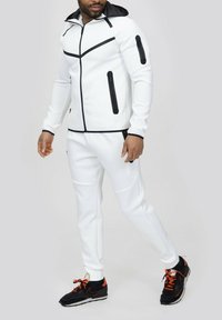 Survêtement de sport blanc avec zips noirs et accents. Comprend une capuche, deux poches latérales et un tissu texturé. Baskets noires avec lacets orange.