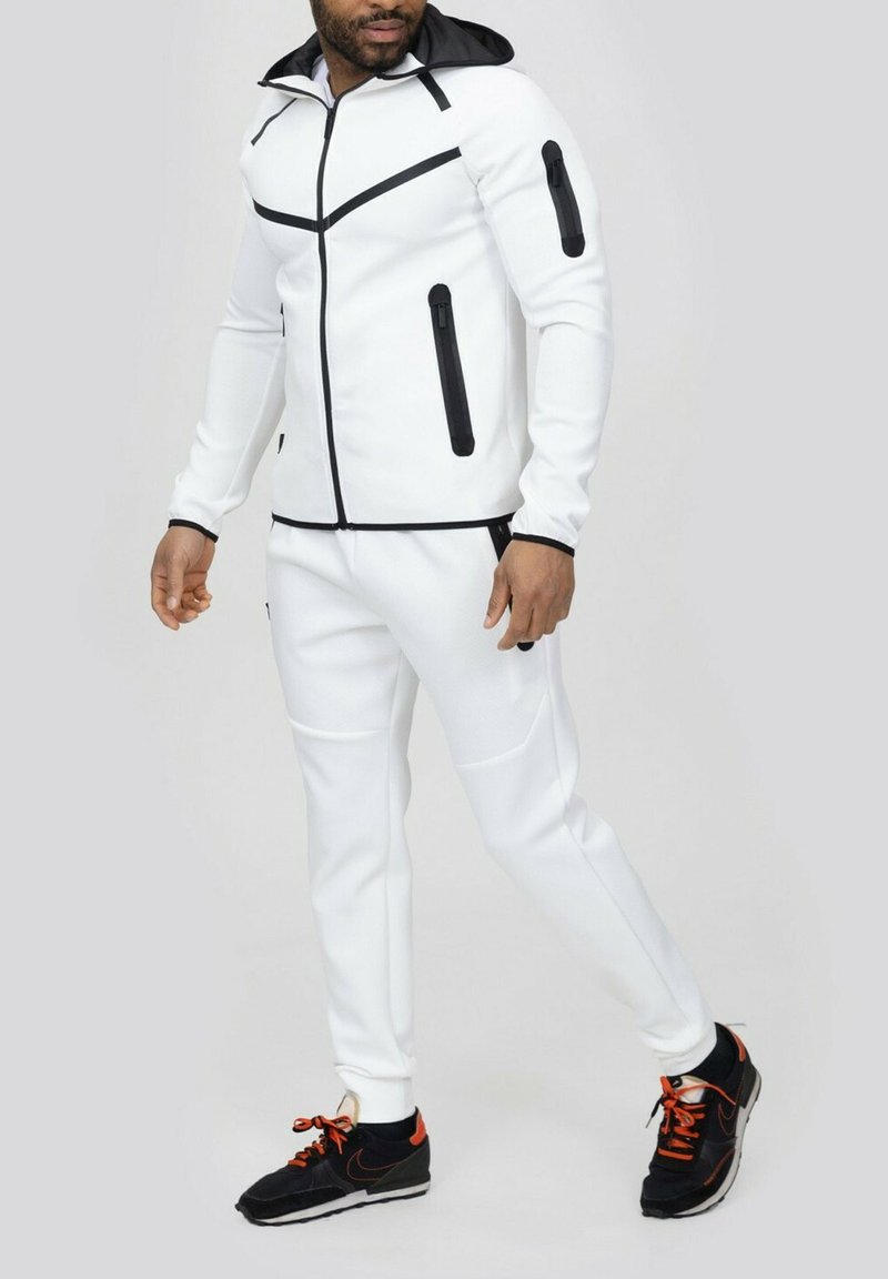 Survêtement de sport blanc avec zips noirs et accents. Comprend une capuche, deux poches latérales et un tissu texturé. Baskets noires avec lacets orange.