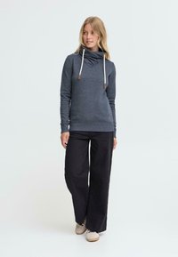 Blå hettegenser med høy krage, med snor og ribbede mansjetter. Kombinert med løse svarte bukser og beige sneakers.