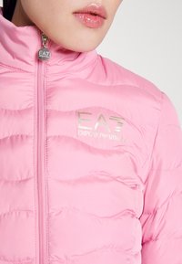 Růžová prošívaná bunda s vodorovným prošíváním, zapínáním na zip a zlatým logem EA7 Emporio Armani na hrudi. Hladký povrch.