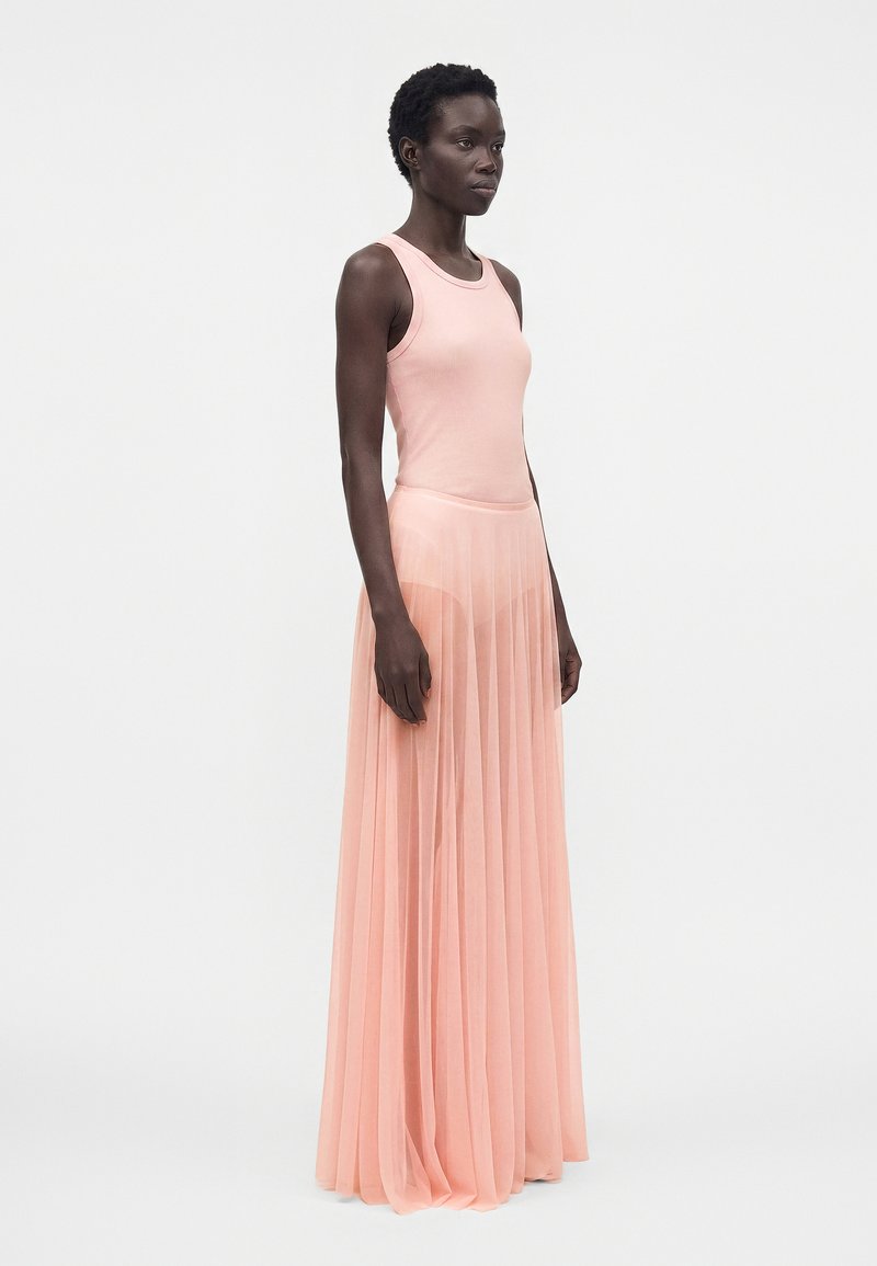 Femme debout, portant un débardeur rose sans manches et une longue jupe plissée transparente par-dessus un short assorti, sur un fond blanc uni.