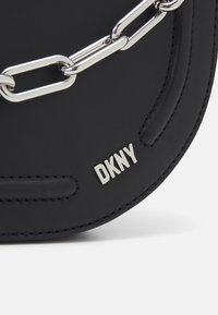 Sac en cuir noir avec une chaîne argentée, des coutures embossées et un logo DKNY métallique à l'avant. Forme arrondie et texture lisse.