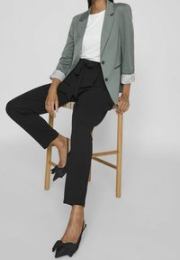 Kobieta siedząca na drewnianym stołku, ubrana w zieloną marynarkę, białą bluzkę, czarne spodnie związywane oraz czarne buty na obcasie z kokardkami.