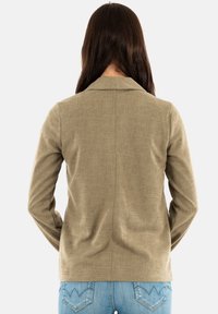 Chaqueta texturizada verde oliva con cuello, mangas largas y detalle en la costura trasera, combinada con jeans de mezclilla azul claro.