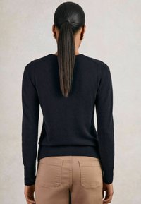Donna con una lunga coda di cavallo, veste un maglione nero e pantaloni beige, in piedi con la schiena rivolta verso la fotocamera su uno sfondo chiaro e uniforme.