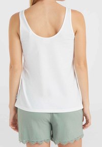 Kobieta w białym, bezrękawnikowym topie i jasnych zielonych szortach z falbankami, widziana od tyłu na tle jasnego, gładkiego tła.