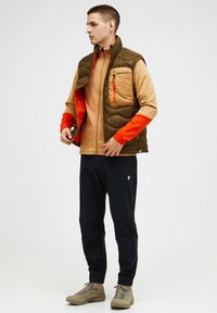 Braune gesteppte Weste mit einer braunen Tasche, orangefarbener Fütterung und langen Ärmeln. Getragen mit einem hellbraunen Oberteil, dunklen Hosen und beigen Schuhen.