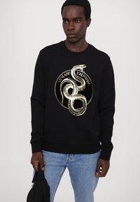 Felpa nera con grafica di un serpente dorato e testo "Armani Exchange". Presenta un colletto tondo classico e polsini a costine. Jeans in denim visibili.