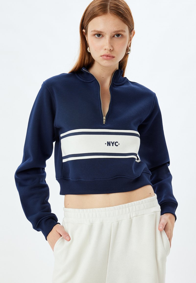 Sudadera recortada de color azul marino con media cremallera, rayas horizontales blancas y un gráfico de "NYC" en la parte delantera. Presenta puños acanalados y una textura suave.