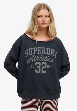 Ung kvinde med langt blondt hår iført en løs sort sweatshirt med teksten "SUPERDRY Athletics No. 32 DPT" og stribede bukser, står roligt.