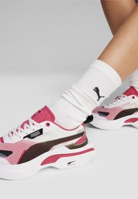 Trampki Puma w kolorze różowym, białym i czarnym z teksturowaną cholewką, geometrycznymi wzorami i grubymi białymi podeszwami; zestawione z prążkowanymi białymi skarpetami.