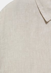Beiger Leinenstoff mit sichtbarer Webstruktur und spitzer Kragenecke, die feine Nahtdetails am Rand zeigt.