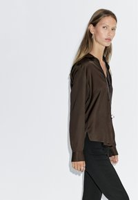 Braune Satinbluse mit Kragen, Knopfleiste und langen Ärmeln. Kombiniert mit schwarzen Hosen, die eine glatte, elegante Textur aufweisen.