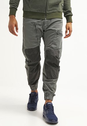 Uomo che indossa pantaloni jogger cargo grigi con rinforzi sulle ginocchia e scarpe da ginnastica blu scuro, in piedi con le mani rilassate lungo i fianchi.