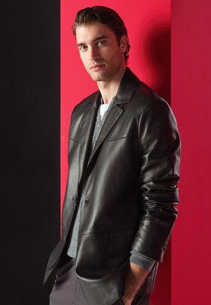 Jonge man met bruin haar, gekleed in een zwarte leren jas en een grijs shirt, staat met handen in de zakken tegen een rood-zwarte achtergrond.