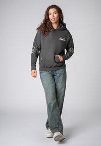 Grijze hoodie met een buidelzak en grafische tekst. Bevat gegraffiteerde elleboogpatches en is gecombineerd met lichtblauwe denim jeans en witte sneakers.