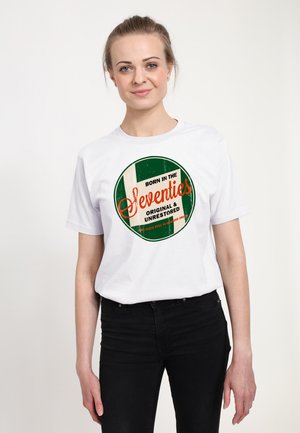 Kvinde iført en hvid T-shirt med en grøn og orange cirkel-logo, der lyder "Født i halvfjerdserne, Original & Urestaureret."