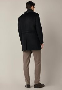 Czarny wełniany płaszcz z wysokim kołnierzem, wyraźną konstrukcją i charakterystycznym szwem z tyłu. Połączony z beżowymi spodniami i ciemnymi butami.