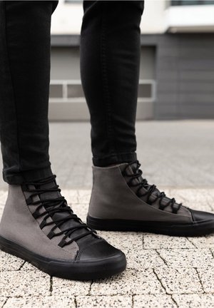 Personne portant un pantalon noir et des baskets montantes grises debout sur un trottoir extérieur texturé.