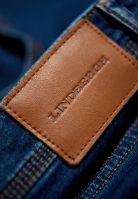 Ciemnoniebieski dżins z prostokątną, brązową skórzaną łatą wypaloną napisem "LINDBERGH", z kontrastowym ściegiem wzdłuż krawędzi.