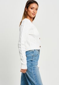 Gilet en maille blanche à col V au design court, avec des poignets et un ourlet côtelés, et des boutons en bois. Associe à un jean bleu taille haute.