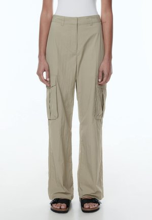 Beige Cargo-Hosen mit lockerer Passform, ausgestattet mit Seitentaschen und einem glatten, leichten Stoff. Getragen mit schwarzen Sandalen.