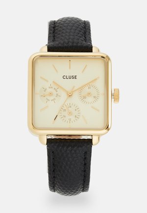 Relógio quadrado em tom dourado com mostrador bege, pulseira de couro preto texturizado, inclui três submostradores que mostram os dias e o formato de 24 horas.