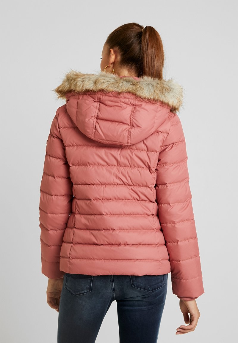 Femme avec queue de cheval brune portant une veste matelassée rose avec capuche bordée de fourrure et un jean foncé, se tenant de dos sur un fond gris.
