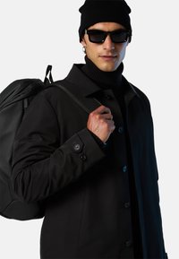 Black waterproof jacket with a buttoned front and flap pockets, paired with a black turtleneck and black backpack. Accessories include a beanie and sunglasses.