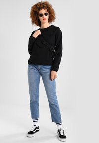 Fekete, hosszú ujjú pulóver fodros részletekkel, világoskék egyenes szárú farmernadrággal és fekete sportcipővel párosítva. A modell napszemüveget visel.