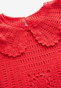 Haut en crochet corail avec un col à volants, présentant un motif de points ouverts et des motifs floraux. Le tissu semble texturé et léger.