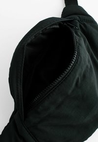 Sac bandoulière noir en tissu avec poche zippée, doté d'une surface texturée et d'une sangle rembourrée pour le confort. L'intérieur est en tissu noir.