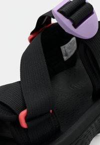 Sandales noires avec des brides texturées, une boucle violette et des accents rouges. La semelle est lisse avec une surface à motifs pour l'adhérence.