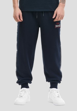 Mann trägt marineblaue Jogginghosen mit weißen Seitenstreifen, weiße Sneakers und ein schwarzes Shirt, steht vor einem hellgrauen Hintergrund.