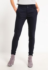 Námořnické kalhoty ve slim fit designu s dvěma předními kapsami, knoflíkovým zapínáním a hladkým povrchem. Spárované s šedými vysokými botami.