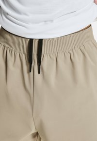 Beżowe spodnie z elastycznym paskiem w talii i czarnym sznurkiem. Gładki materiał w luźnym kroju, z bocznymi szwami i bez widocznych kieszeni.