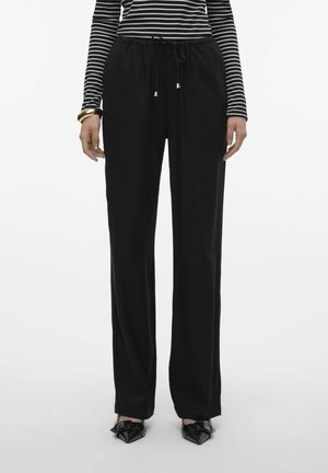 Pantaloni neri larghi con vita regolabile a cordoncino, dettagli in metallo dorato e una texture liscia, abbinati a scarpe nere a punta.