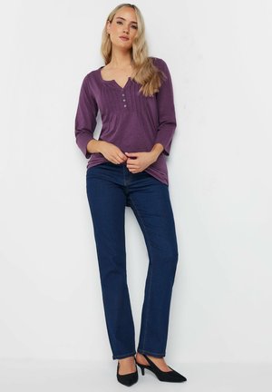 Maglione lungo a maniche con collo rotondo di colore viola, con dettagli a bottone e texture a coste, abbinato a jeans in denim blu scuro e scarpe con tacco nere.