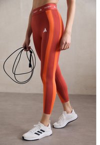 Punane ja oranžid spordileggingud, mis on sobiva disainiga, koos logoga puusal, valgete treeningkingade ja hüppenööriga.