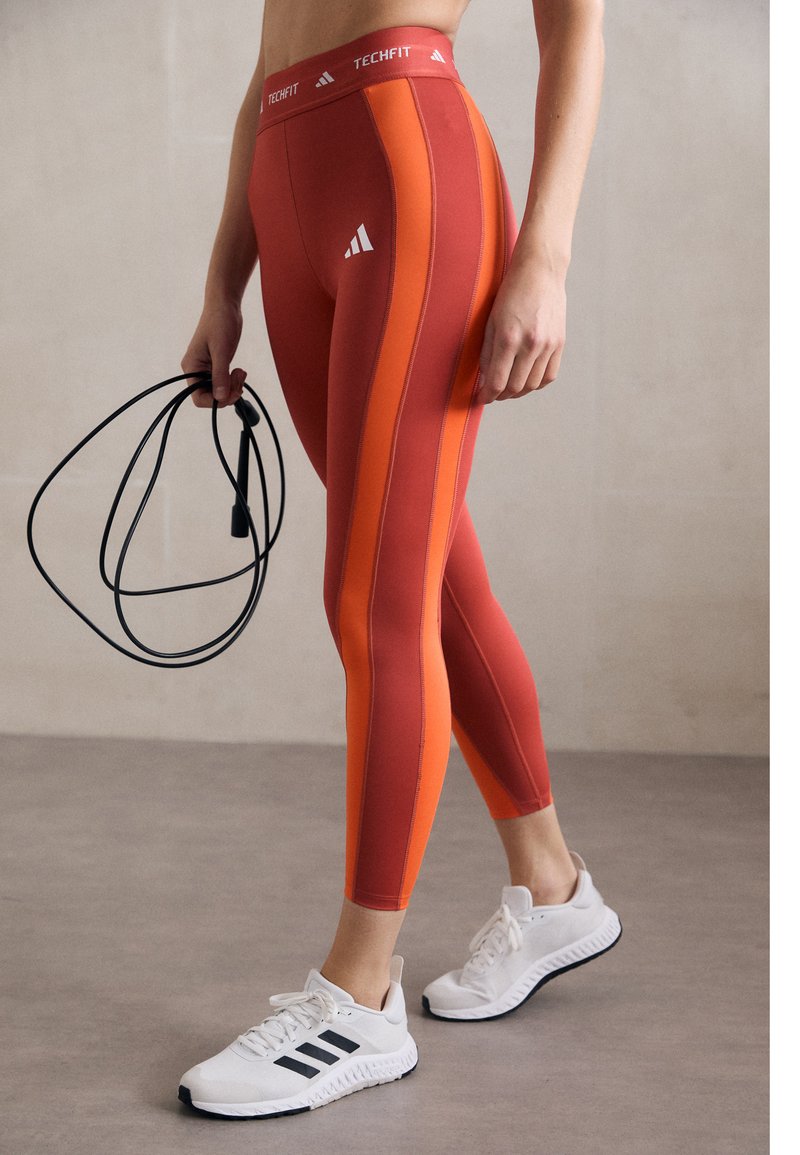 Punane ja oranžid spordileggingud, mis on sobiva disainiga, koos logoga puusal, valgete treeningkingade ja hüppenööriga.