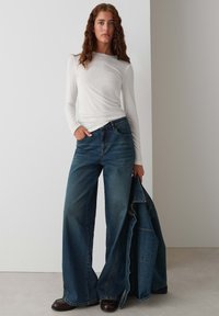 Weißes Langarmshirt, kombiniert mit weitem blauen Denim-Jeans. Das Modell hält eine Jeansjacke. Glatte Stoffstruktur, lässiges Design, entspannte Passform.