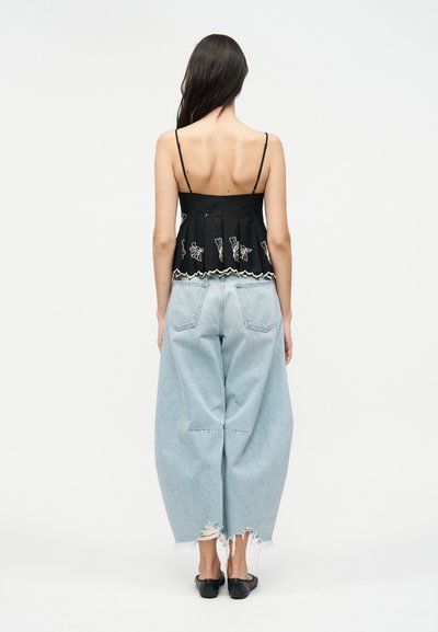 Γυναίκα που στέκεται με την πλάτη προς τα εμπρός φορώντας μαύρο τοπ με λεπτές τιράντες και λευκή κεντημένη λουλουδένια διακόσμηση και φαρδιά, ανοιχτό μπλε τζιν με μαύρα μπαλαρίνια.