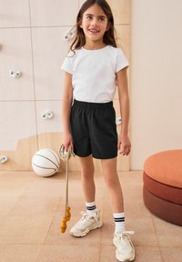 Svarta shorts med elastiskt midjeband, vit t-shirt, vita sneakers med svarta detaljer, håller i ett rep. Mjuk golvbeläggning och en basketboll närvarande.