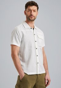 Camisa de lino blanca de manga corta con botones frontales, botones negros y un bolsillo en el pecho. Textura suave y corte casual.