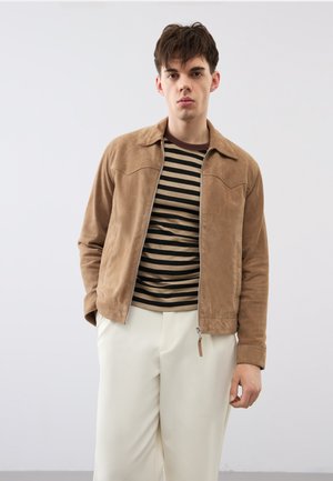 Giovane uomo che indossa una giacca di pelle scamosciata color cuoio sopra una camicia a righe nere e marroni e pantaloni bianco sporco, in piedi contro uno sfondo chiaro e uniforme.