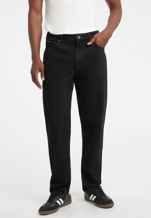 Jeans de mezclilla negra con corte recto, diseño de cinco bolsillos, cierre con botón plateado y detalle de costura minimalista. Combinados con zapatillas negras.