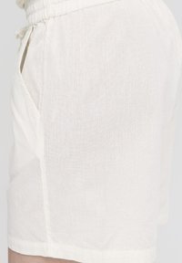 Shorts en lin de couleur crème clair, avec une coupe décontractée, une taille élastique et des poches latérales, offrant une texture lisse.