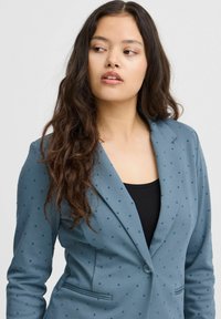 Tealblazer med mörka prickar, strukturerad design, slagkrage, två framfickor och knäppning. Bärs över en svart topp.