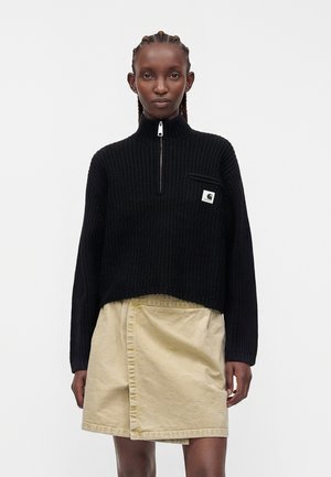 Donna che indossa un maglione nero con mezza cerniera e logo sulla tasca del petto e una gonna beige a portafoglio, in piedi davanti a uno sfondo chiaro e uniforme.